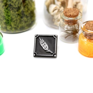 May include: A silver metal pin with a wheat stalk design. The pin is square and has a black background. The wheat stalk is silver and is in the center of the pin.