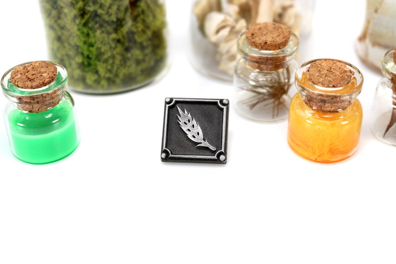 May include: A silver metal pin with a wheat stalk design. The pin is square and has a black background. The wheat stalk is silver and is in the center of the pin.