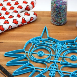 May include: A blue plastic cookie cutter shaped like the iconic Majora's Mask from the Legend of Zelda video game series. The cutter is on a wooden surface with a rolling pin and sprinkles in jars.