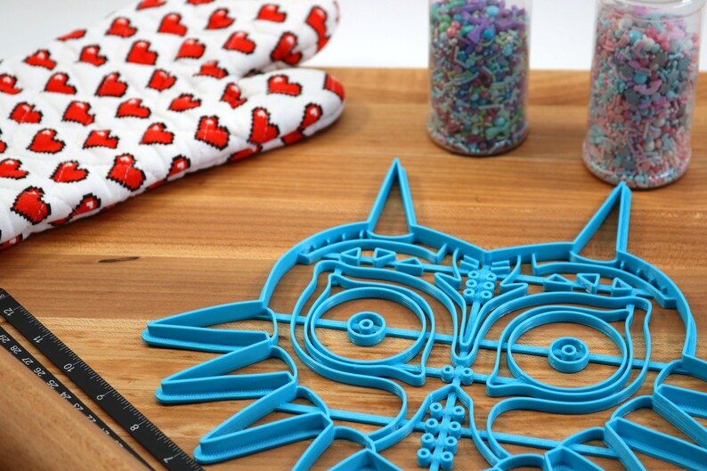 May include: A blue plastic cookie cutter shaped like the iconic Majora's Mask from the Legend of Zelda video game series. The cutter is on a wooden surface with a rolling pin and sprinkles in jars.