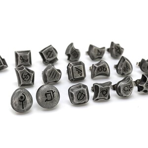 May include: A set of 17 silver-toned metal charms featuring a variety of symbols and designs. The charms are small and have a textured finish.