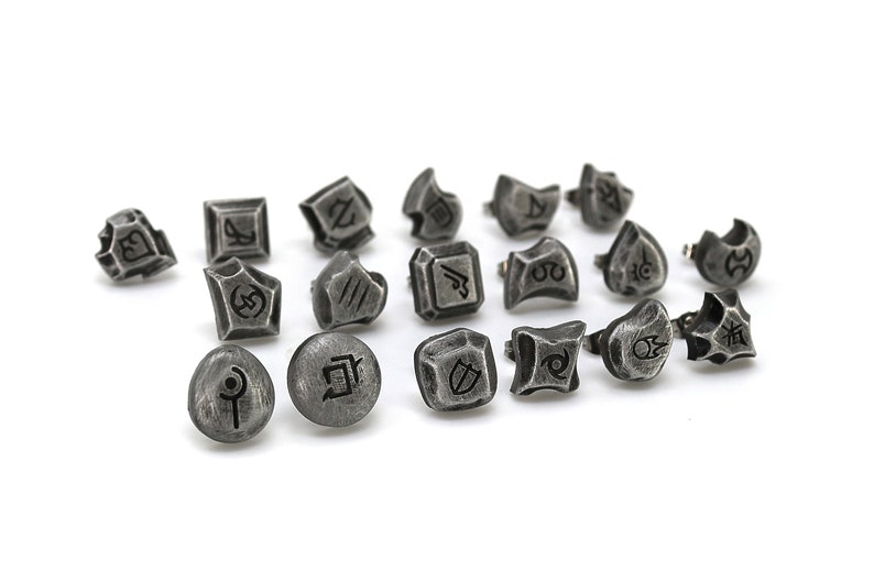 May include: A set of 17 silver-toned metal charms featuring a variety of symbols and designs. The charms are small and have a textured finish.