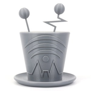 Peut inclure: Un pot de fleurs robot gris imprimé en 3D avec une antenne en zigzag et deux boutons ronds sur le dessus. Le pot a un motif géométrique sur le devant et une base en soucoupe.