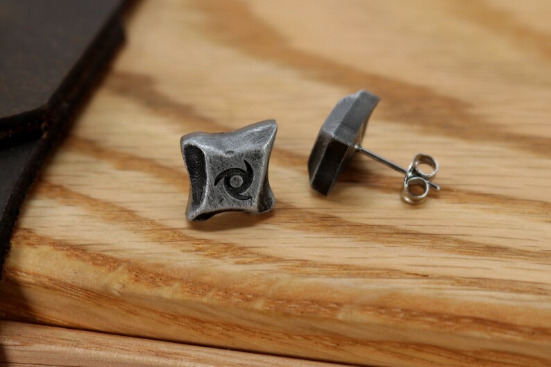 Puede incluir: Un par de pendientes de bot&oacute;n plateados. Cada pendiente tiene un dise&ntilde;o cuadrado y texturizado con un s&iacute;mbolo de espiral en el centro. Los pendientes se muestran sobre una superficie de madera.