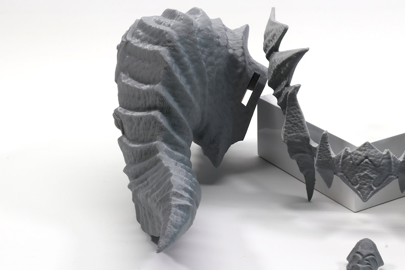 May include: A gray 3D printed dragon head sculpture with detailed scales and horns. The sculpture is broken into multiple pieces and is laying on a white surface.