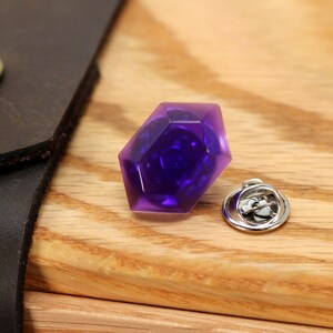 May include: A purple faceted gem pin with a silver pin back. The gem is a dark purple color and has a faceted design.