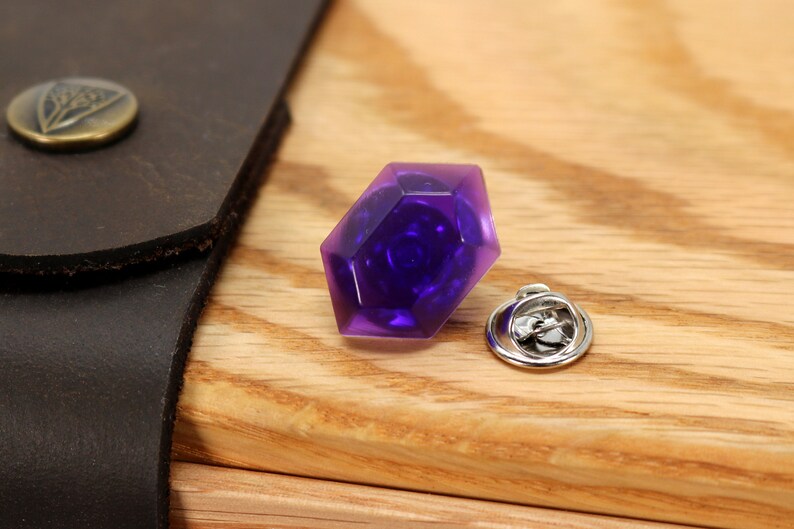 May include: A purple faceted gem pin with a silver pin back. The gem is a dark purple color and has a faceted design.