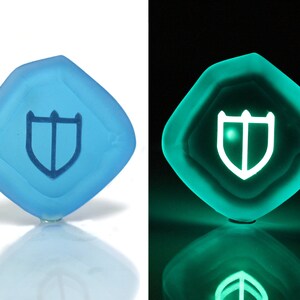 Puede incluir: Un dado de color azul claro en forma de diamante con un s&iacute;mbolo de escudo blanco. El dado brilla en verde en la oscuridad.