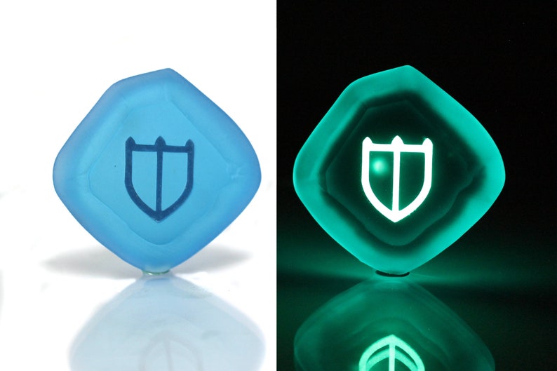 Puede incluir: Un dado de color azul claro en forma de diamante con un s&iacute;mbolo de escudo blanco. El dado brilla en verde en la oscuridad.