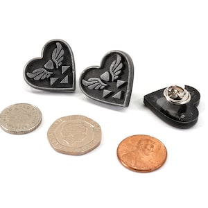 Puede incluir: Dos pines plateados en forma de coraz&oacute;n con un s&iacute;mbolo alado y un pin negro en forma de coraz&oacute;n con un cierre de seguridad.
