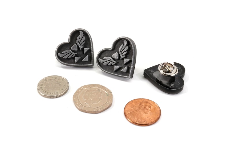 Puede incluir: Dos pines plateados en forma de coraz&oacute;n con un s&iacute;mbolo alado y un pin negro en forma de coraz&oacute;n con un cierre de seguridad.