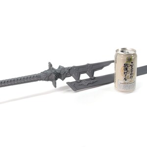 May include: A grey sword with a decorative handle and a matching scabbard. The sword is lying on a white surface next to a can of Japanese beer.