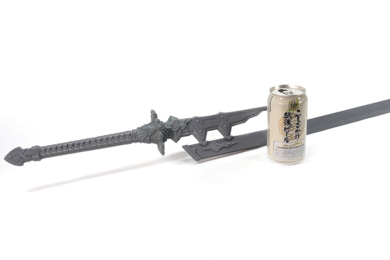 May include: A grey sword with a decorative handle and a matching scabbard. The sword is lying on a white surface next to a can of Japanese beer.