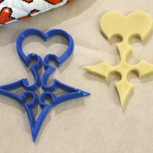 May include: A blue plastic cookie cutter shaped like a heart with a star in the center. A plain, light brown cookie is cut out using the cookie cutter.
