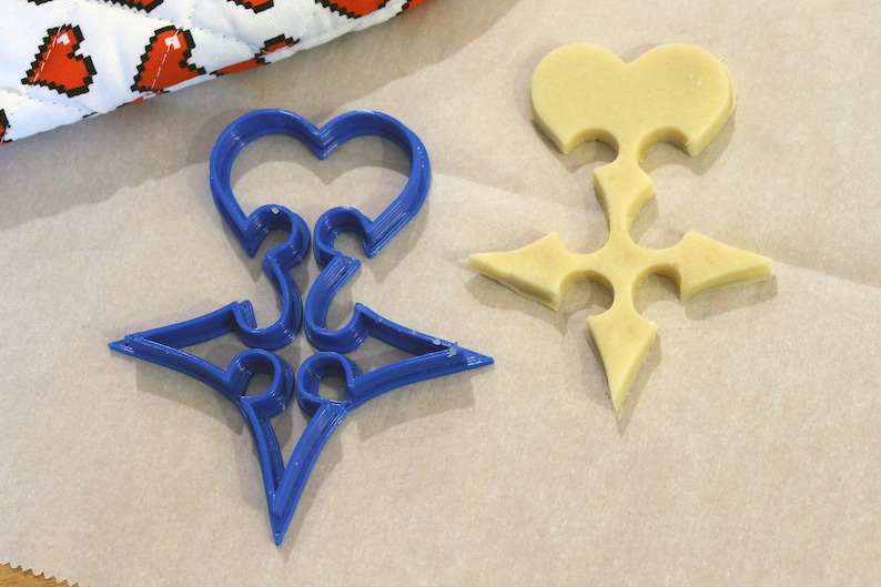 May include: A blue plastic cookie cutter shaped like a heart with a star in the center. A plain, light brown cookie is cut out using the cookie cutter.
