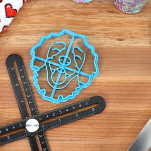 May include: A blue plastic cookie cutter shaped like a dog's face. The cutter is on a wooden surface with a black ruler and a blue spatula.