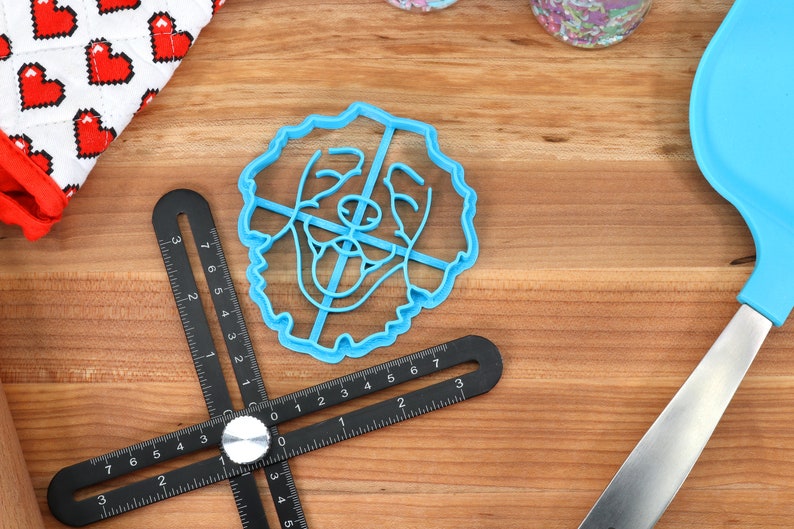 May include: A blue plastic cookie cutter shaped like a dog's face. The cutter is on a wooden surface with a black ruler and a blue spatula.