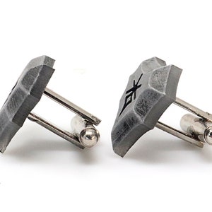 May include: A pair of silver cufflinks with gray, textured, square-shaped tops. Each top has a black symbol on it.
