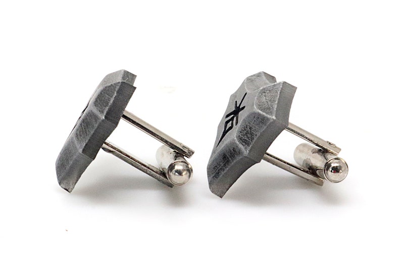 May include: A pair of silver cufflinks with gray, textured, square-shaped tops. Each top has a black symbol on it.