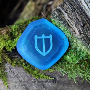 Puede incluir: Una piedra preciosa de vidrio azul con un dise&ntilde;o de escudo. La piedra preciosa est&aacute; sentada en un tronco cubierto de musgo.