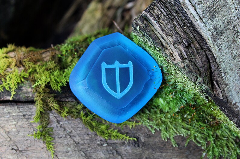 Puede incluir: Una piedra preciosa de vidrio azul con un dise&ntilde;o de escudo. La piedra preciosa est&aacute; sentada en un tronco cubierto de musgo.