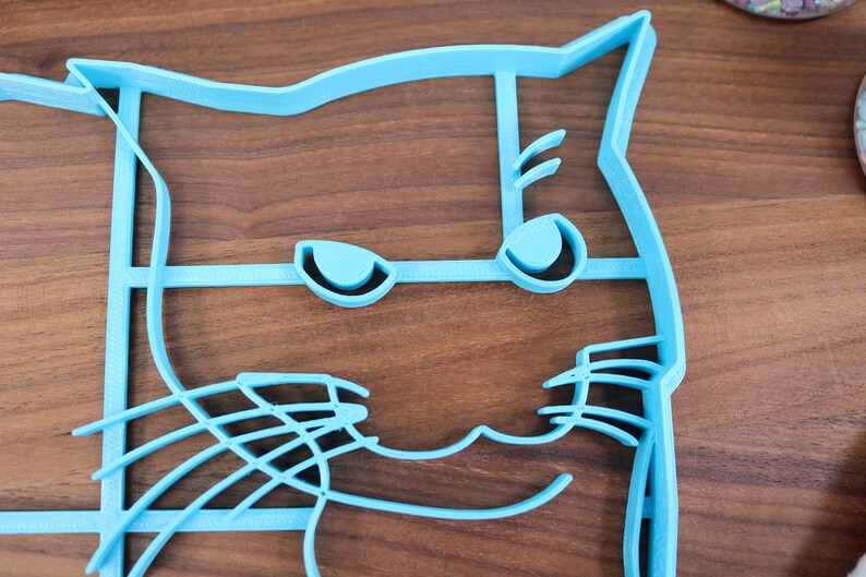 May include: A light blue plastic cat-shaped cookie cutter with a detailed design of eyes, whiskers, and a mouth.