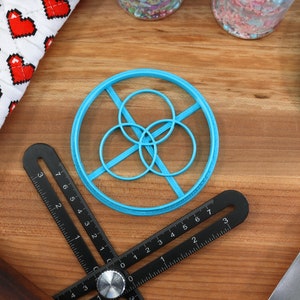 May include: A blue plastic cookie cutter shaped like a circle with three smaller circles inside. The cutter is on a wooden surface with a black ruler and a blue spatula.