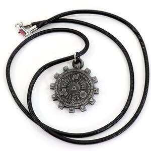 May include: A silver-colored pendant necklace with a circular design featuring a stylized Triforce symbol and other geometric patterns. The necklace is on a black cord.
