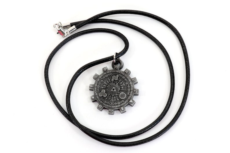 May include: A silver-colored pendant necklace with a circular design featuring a stylized Triforce symbol and other geometric patterns. The necklace is on a black cord.