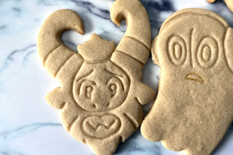 K&ouml;nnte beinhalten: Zwei Zuckerkekse, die mit Zuckerguss verziert sind. Ein Keks ist in Form eines Geistes mit zwei gro&szlig;en Augen und einem kleinen Mund. Der andere Keks ist in Form eines Monsters mit H&ouml;rnern, gro&szlig;en Augen und einem gro&szlig;en Mund.