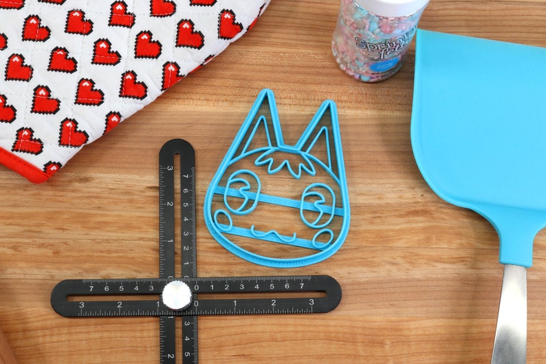 May include: Blue plastic cookie cutter shaped like a cartoon cat with large eyes and a wide smile. The cutter is on a wooden surface with a ruler and a spatula.