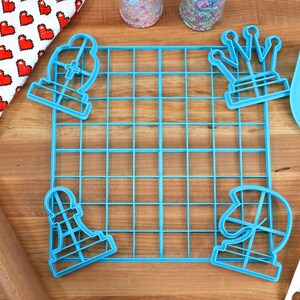 May include: Blue plastic chess piece cookie cutters, a blue grid baking mat, a wooden rolling pin, a blue spatula, and two jars of sprinkles on a wooden surface.