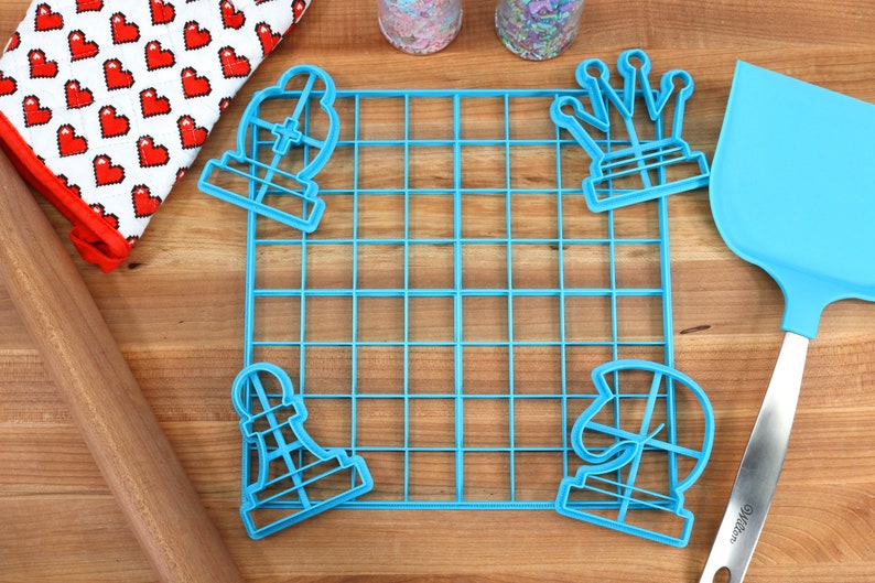 May include: Blue plastic chess piece cookie cutters, a blue grid baking mat, a wooden rolling pin, a blue spatula, and two jars of sprinkles on a wooden surface.