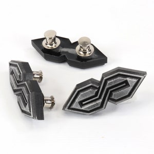 May include: Three geometric-shaped lapel pins with silver-colored posts. The pins are dark gray with a textured, metallic appearance. The design features a stylized, repeating pattern. The pins are displayed on a white surface.