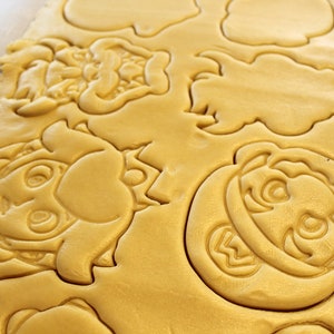 May include: A sheet of unbaked cookie dough with various cartoon character shapes cut out. The shapes include a mushroom, a character with a mustache, and a character with a crown.