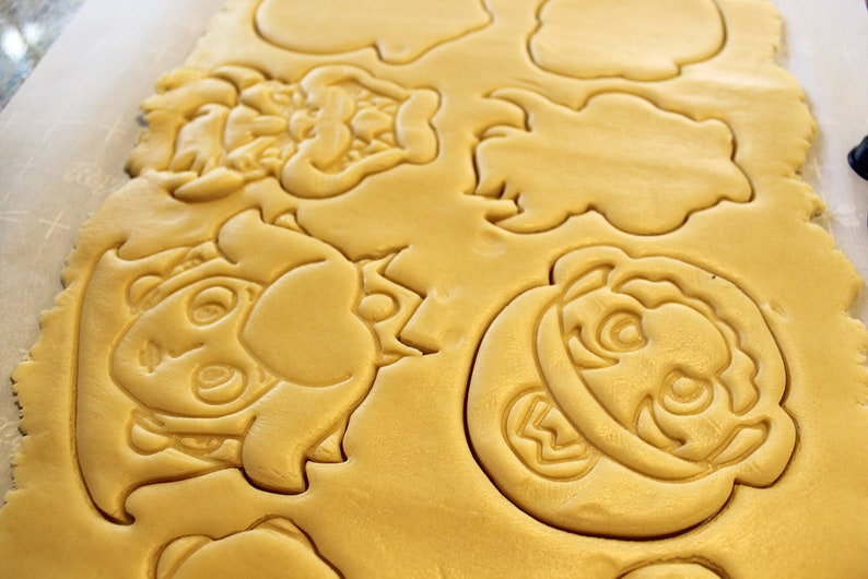 May include: A sheet of unbaked cookie dough with various cartoon character shapes cut out. The shapes include a mushroom, a character with a mustache, and a character with a crown.