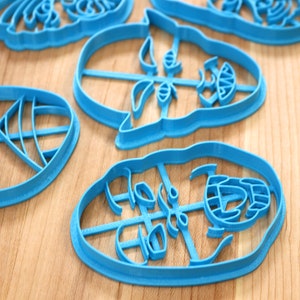 May include: Four blue plastic cookie cutters shaped like faces. The cutters are on a wooden surface.