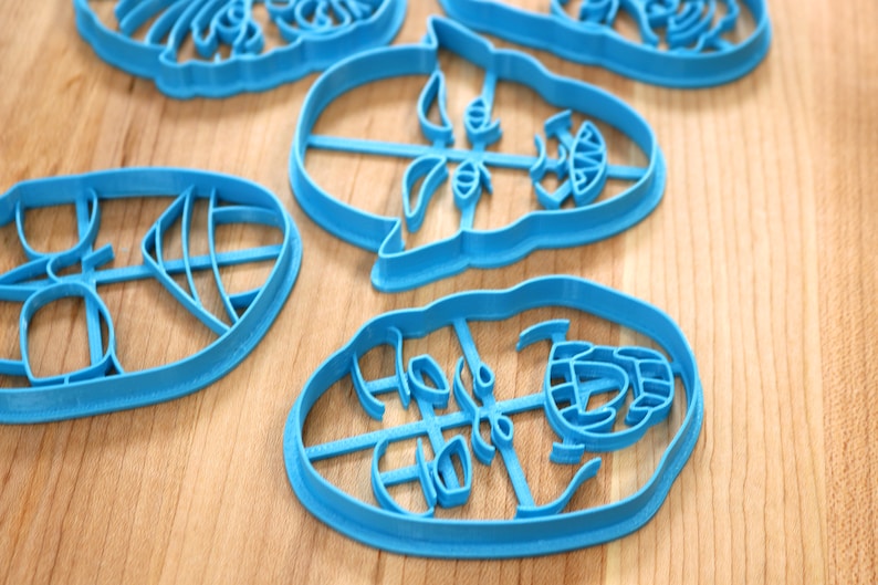 May include: Four blue plastic cookie cutters shaped like faces. The cutters are on a wooden surface.