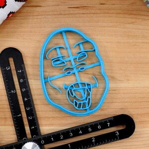 May include: A blue plastic cookie cutter shaped like a face with a wide open mouth and teeth. The face has a surprised expression with wide eyes and raised eyebrows.