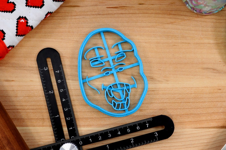 May include: A blue plastic cookie cutter shaped like a face with a wide open mouth and teeth. The face has a surprised expression with wide eyes and raised eyebrows.