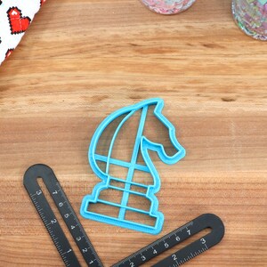May include: A blue plastic cookie cutter shaped like a chess knight. The cutter is on a wooden surface with a ruler and other baking supplies.