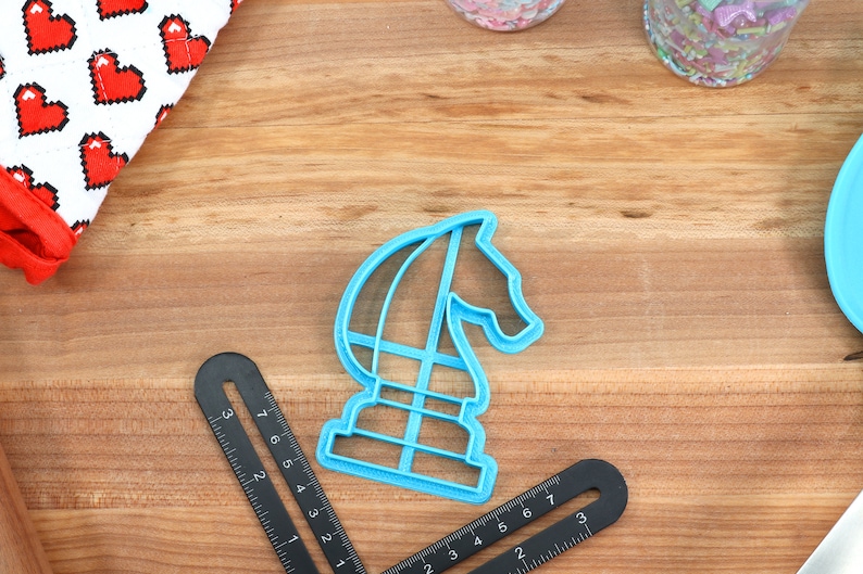 May include: A blue plastic cookie cutter shaped like a chess knight. The cutter is on a wooden surface with a ruler and other baking supplies.