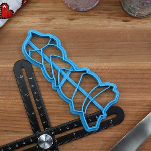 K&ouml;nnte beinhalten: Ein blauer Pl&auml;tzchenausstecher aus Kunststoff in Form eines Stapels von drei Weihnachtsglocken. Der Ausstecher liegt auf einer Holzoberfl&auml;che mit einem Lineal und einem blauen Spatel.