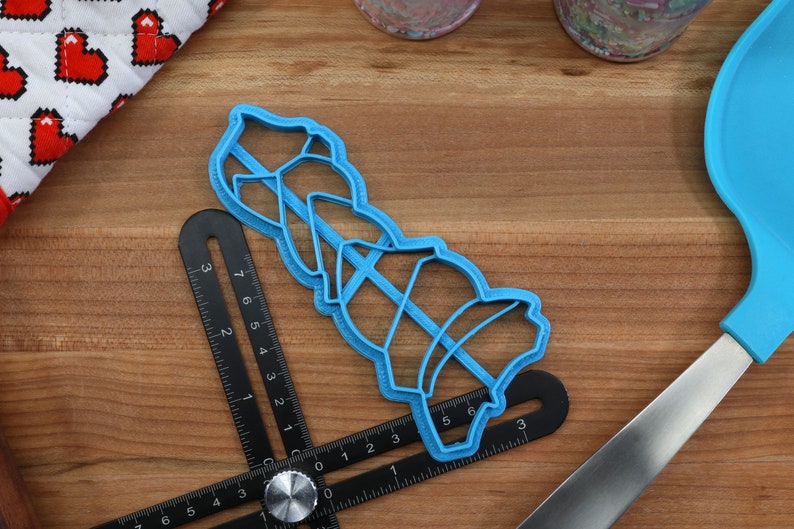 K&ouml;nnte beinhalten: Ein blauer Pl&auml;tzchenausstecher aus Kunststoff in Form eines Stapels von drei Weihnachtsglocken. Der Ausstecher liegt auf einer Holzoberfl&auml;che mit einem Lineal und einem blauen Spatel.