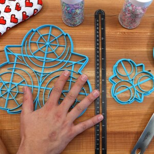 May include: A set of three blue plastic cookie cutters, one large circle with a web design, one smaller circle with a web design, and a three-circle flower design.