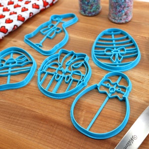 May include: Five blue plastic cookie cutters shaped like Easter eggs, a bunny, and a bunny in an egg. The cutters are on a wooden surface.