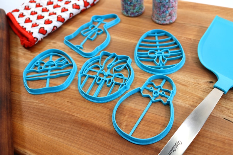 May include: Five blue plastic cookie cutters shaped like Easter eggs, a bunny, and a bunny in an egg. The cutters are on a wooden surface.