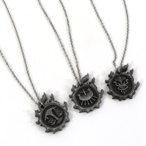 May include: Three silver necklaces with pendants featuring different symbols. Each pendant is a circular design with a black and silver finish. The symbols are a ship, a tree, and a flower.