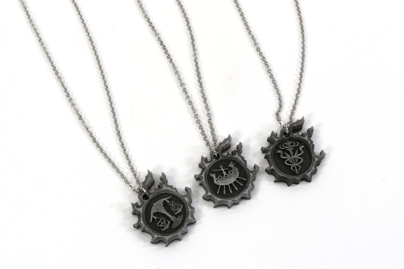May include: Three silver necklaces with pendants featuring different symbols. Each pendant is a circular design with a black and silver finish. The symbols are a ship, a tree, and a flower.