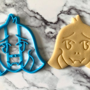 May include: A blue plastic cookie cutter shaped like a cartoon character with a sad expression, next to a light brown cookie with the same design.